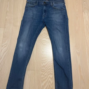 Blå Luke jeans från Lee W31 L30 - Snygga blå jeans från Lee, modell Luke, med klassisk femficksdesign och smal passform. Jeansen har en mörk tvätt med lätt slitning och raka ben. Tillverkade i mjukt bomullsmaterial med lite stretch för extra komfort.
