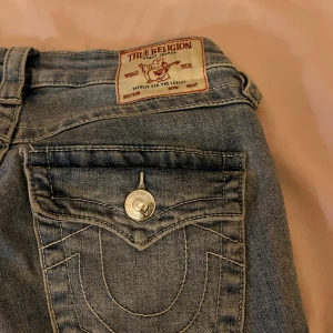 True Religion Becca Bootcut Jeans - Snygga blå jeans från True Religion, modell Becca med bootcut och midwaist. Klassiska fickor med dekorativa sömmar och silverfärgade knappar. Tillverkade i jeansmaterial med coola detaljer på bakfickorna. Perfekt för dig som gillar en avslappnad men trendig look. Jag har använt dem en gång! men bara i någon timme då jag märkte att de kändes för stora. Jag är 1,58 cm lång. Priset går att diskutera!☺️