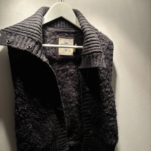 Mörkgrå väst från H&M L.O.G.G. - Mörkgrå väst med grov struktur och bred ribbad krage. Västen har knappar framtill och är tillverkad i ett mjukt, ulligt material som ger en chunky känsla. Perfekt för lager-på-lager och chill stil. 62% ull och 24% polyamid