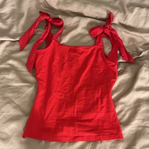 Röd linnetopp med knyt från Lindex - Snygg röd linnetopp från Holly & Whyte by Lindex i storlek XS. Toppen har breda axelband med knyt som ger en söt och trendig look. Tillverkad i mjuk bomull och perfekt för varma dagar. Enkel, figurnära passform och stilren design.