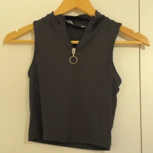 Svart ribbad croppad linnetopp - Säljer en svart croppad linnetopp med ribbad struktur och en cool dragkedja framtill med rund metallring. Toppen är ärmlös och har en hög krage som ger en edgy vibe. Perfekt att styla med jeans eller kjol för en trendig look.