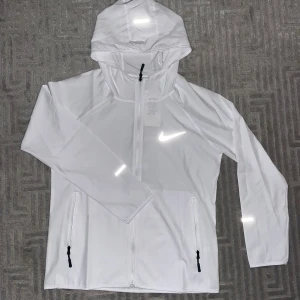 Vit vindjacka med huva från Nike 160-170cm - Snygg vit vindjacka från Nike med huva och hel dragkedja. Jackan har reflekterande detaljer, två fickor med dragkedja och sportig design. Perfekt för träning och aktiva dagar. 