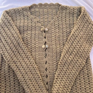 Beige virkad kofta med pärlknappar - Superfin beige virkad kofta med lång ärm och snygga pärlknappar framtill. Koftan har ett unikt virkat mönster och vågig kant längs halsringningen. Perfekt att slänga över en topp för en chill och trendig look.