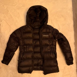 Svart Peak Performance dunjacka - Säljer en svart dunjacka från Peak Performance med huva och dragkedja. Jackan har en puffad look, två sidofickor med dragkedja och diskret logga på bröstet. Perfekt för kalla dagar och har en clean, modern stil som funkar till det mesta. Kolla sista bilden för ett pyttelitet hål som dessutom är lagat! Inget man lägger märke till men bör nämnas :) köptes för 4200kr! Använd fåtal gånger 