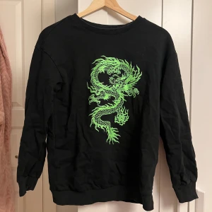 Svart tröja med grön drake från SHEIN - Cool svart tröja från SHEIN med en stor grön drake tryckt på framsidan. Tröjan har rund hals, långa ärmar och ribbade muddar. Perfekt för dig som vill sticka ut med en unik och edgy stil.