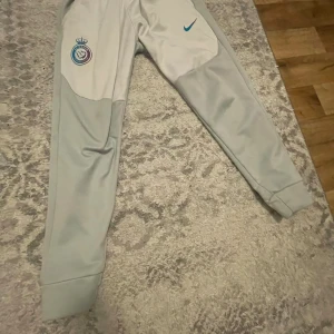 Nike Alnasser byxor  - Nike träningsbyxor i ljusgrått med klubbmärke och blå Nike-logga på framsidan. Slimmad passform med muddar vid benslut och elastisk midja. Perfekta för sport eller chill, tillverkade i mjukt syntetmaterial som andas.