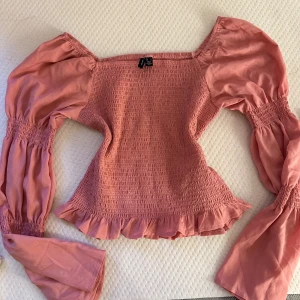 Rosa blus med puffärmar  - Supersöt rosa blus  med smockad kropp och volangkant nertill. Blusen har långa puffärmar med smock vid ärmslut och en fyrkantig halsringning. Perfekt för dig som gillar romantisk och trendig stil.