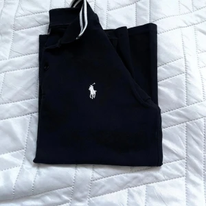 Svart pikétröja från Ralph Lauren - Klassisk svart pikétröja från Ralph Lauren med vit broderad logga på bröstet. Tröjan har korta ärmar, krage med vita ränder och knäppning framtill. Perfekt för en clean och sportig stil. Tillverkad i mjuk bomull som andas. Passar personer som är ca 150-160cm långa. Finns en spricka, kom privat för bilder!