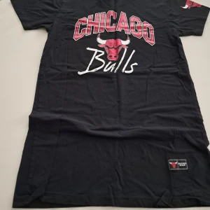 Chicago Bulls svart t-shirt XS - Svart t-shirt med Chicago Bulls tryck i rött och vitt framtill. Klassisk passform med korta ärmar och rund hals. Tillverkad i mjuk bomull, perfekt för dig som gillar basket och streetwear. Officiell NBA-logga på insidan och Bulls-märke nertill.