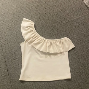 Vit oneshoulder topp med volang - Säljer en vit oneshoulder topp med volang, passar bra både till vardags och till fester, använt endast 2-3 gånger, mycket bra kvalitet.