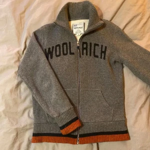 Grå kofta från Woolrich med dragkedja - Storlek L men passar mer S kanske MSnygg grå kofta från Woolrich med svart logotyp på bröstet. Tröjan har ribbad hög krage, muddar och nederkant med orange och svarta ränder. Praktiska fickor framtill och hel dragkedja. Perfekt för kyliga dagar och riktigt chill stil.