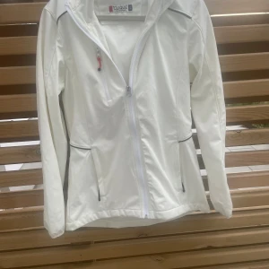 Vit vindjacka från Clique, storlek S - Snygg vit vindjacka från Clique i storlek S. Jackan har hel dragkedja framtill, två sidofickor med dragkedja och en bröstficka med röd detalj. Långärmad och tillverkad i ett slätt syntetmaterial, perfekt för blåsiga dagar.