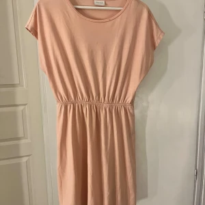Ljusrosa kort klänning från Vila - Superfin ljusrosa kort klänning från Vila med rund halsringning och resår i midjan. Klänningen har korta ärmar och är gjord i mjuk bomull, perfekt för varma dagar. Enkel och stilren design som är lätt att matcha med sneakers eller sandaler.