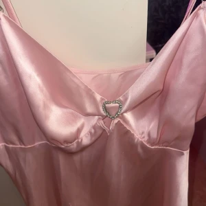 Ljusrosa satin klänning med hjärtdetalj - Supersöt ljusrosa klänning i satin med tunna axelband och en glittrande hjärtformad detalj framtill. Klänningen har en mjuk, glansig yta och en feminin siluett som ger en romantisk vibe. Perfekt för dig som älskar gulliga detaljer och en chic look.