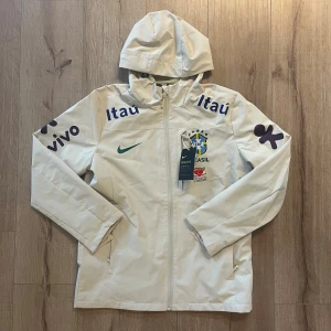 Vit Nike Brasil vindjacka med huva - Snygg vit vindjacka från Nike med Brasil-logga och sponsormärken på bröst och ärmar. Jackan har huva, hel dragkedja och två sidofickor. Perfekt för dig som gillar fotboll och vill ha en sportig stil. Materialet är lätt och känns fräscht.