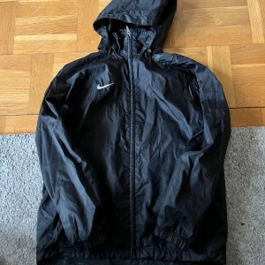 Svart Nike vindjacka med huva - Svart vindjacka från Nike med huva och vit logga på bröstet. Jackan har meshfoder, dragkedja framtill, snörning i nederkant och praktiska fickor med tryckknappar. Perfekt för blåsiga dagar och sportiga outfits.