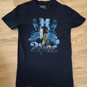 Svart 2Pac t-shirt från Mister Tee - Svart t-shirt från Mister Tee med stort 2Pac-tryck på framsidan. Motivet har coola blå moln och en stor 2Pac-text i silvergrått. Klassisk rund hals och korta ärmar, perfekt för dig som gillar streetwear och hiphop.