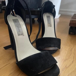 Svarta mocka-klackar från NLY Shoes - Snygga svarta klackar från NLY Shoes med bred klack och öppen tå. Skorna har en stilren design med mocka-material och smala remmar som går runt ankeln för extra support. Perfekta för dig som vill ha en chic och modern look. Jag har klistrat på gelsulor framtill i skon för extra komfort. I okej skick, använt ett fåtal gånger. 
