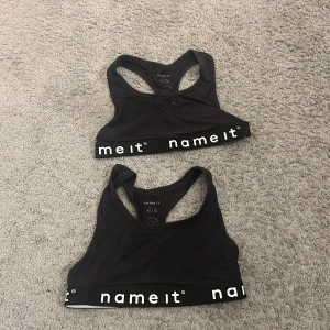 Svarta sportiga toppar från Name It - Två svarta sportiga toppar från Name It med bred resår nertill där det står 'name it' i vitt. Modellen är ärmlös med brottarrygg och tillverkad i mjuk bomull. Perfekta för träning eller chill dagar.