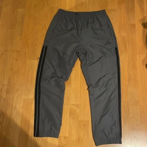 Grå träningsbyxor från Adidas - Snygga grå träningsbyxor från Adidas med svarta ränder längs sidorna och elastisk midja. Byxorna har en rak passform och är tillverkade i ett lätt syntetmaterial som passar perfekt för sport och aktivitet. Diskret Adidas-logga på framsidan. I princip oanvända, är i storlek M men kan passa som L