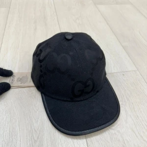 Gucci jumbo svart  - Snygg svart keps från Gucci med GG-logga i ton-i-ton över hela kepsen. Justerbar rem baktill i skinn och klassisk böjd skärm. Materialet är slitstark bomull och kepsen har en cool, mörk vibe som passar perfekt till streetwear.