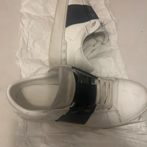 Valentino Garavani vita sneakers strl 43 - Snygga vita Valentino Garavani sneakers med bred svart rem över snörningen och guldtext på plösen. Skorna har platt sula, rund tå och är tillverkade i skinn. Perfekt för dig som gillar exklusiva och stilrena sneakers med unik design.