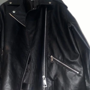 Svart skinnjacka från Missguided - Säljer en svart skinnjacka från Missguided i klassisk bikerstil med silvriga dragkedjor och detaljer. Jackan har bred krage, snedställda fickor med dragkedja och coola metallknappar. Perfekt för dig som vill ha en edgy look.