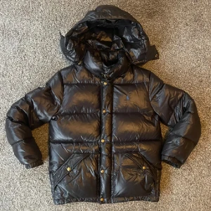 Svart dunjacka från Polo Ralph Lauren - Säljer en svart, glansig dunjacka från Polo Ralph Lauren med huva och guldiga knappar. Jackan har klassisk pufferstil, två stora fickor framtill och broderad blå logga på bröstet. Perfekt för kalla dagar och riktigt snygg till jeans. Pris kan diskuteras 