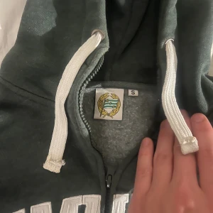 Mörkgrön Hammarby hoodie - Säljer en mörkgrön hoodie från Hammarby med vit broderad text 'HAMMARBY SÖDERMALM' på bröstet och klubbmärke på ärmen. Litet Slitage som ni ser