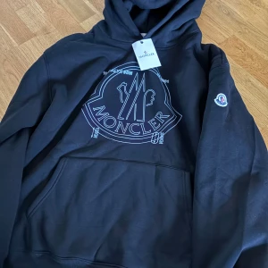 Svart hoodie från Moncler XL - Svart hoodie från Moncler med stor logga framtill och klassisk känguruficka. Tröjan har huva och Moncler-märke på ärmen. Perfekt för dig som gillar streetwear och vill ha något snyggt och bekvämt.