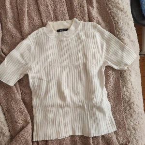Vit ribbad topp från Gina Tricot - Säljer en vit ribbad topp från Gina Tricot i storlek S. Toppen har korta ärmar, rund hals och ett snyggt vertikalt ribbat mönster som ger en clean och fräsch look. Perfekt att styla med jeans eller kjol för en enkel men trendig outfit.