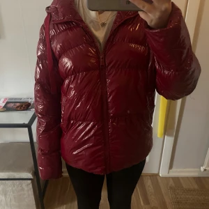 Vinröd pufferjacka från Guess - Säljer en glansig vinröd pufferjacka från Guess med huva och breda snören med logga. Du behöver denna coola jacka i ❄️. Bara använd några gånger! Hittar ingen storlek men för referens är jag 165cm lång och har vanligtvis Xs/S