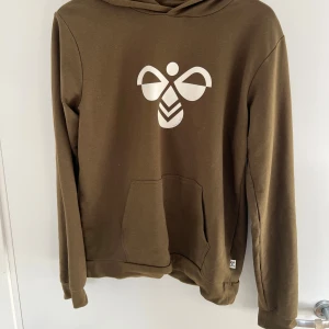 Olivgrön hoodie från Hummel - Snygg olivgrön hoodie från Hummel med deras klassiska bi-logga i vitt på bröstet. Tröjan har huva, magficka och långa ärmar. Perfekt för dig som gillar en avslappnad och sportig stil.
