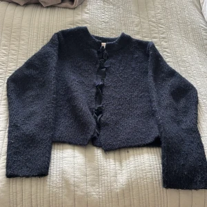 Marinblå kofta med rosetter från Gina Tricot - Mysig marinblå kofta från Gina Tricot i stickat material. Koftan har långa ärmar och stängs framtill med flera söta rosetter. Perfekt för lager-på-lager och ger en cool touch till din outfit.