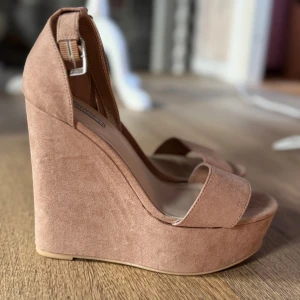 Ljusrosa  platåsandaler från NLY Shoes - Säljer ett par snygga klackar med hög platå och kilklack från NLY Shoes. Skorna har öppen tå, bred rem över foten och justerbart spänne vid ankeln. Ytan är i mockaimitation som ger en mjuk känsla och trendig look.