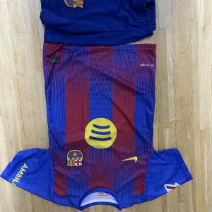Barcelona fotbollströja Yamal #10 - Säljer en Barcelona fotbollströja med Yamal #10 på ryggen. Tröjan är kortärmad och har klassiska blå och röda vertikala ränder, gula detaljer och klubbmärke på bröstet. Materialet är lätt och andas, perfekt för match eller träning.