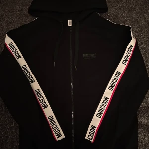 Svart Moschino Kofta - Svart Kofta från Moschino med huva och dragkedja framtill. Längs ärmarna finns vita band med Moschino-logga och röda/svarta detaljer. Köpt för cirka 3000kr säljer för 1599kr, pris går att diskuteras vid snabb affär