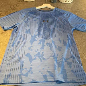 Blå compression shirt  från Under Armour - Snygg blå t-shirt från Under Armour med coolt geometriskt mönster och diskret camo-detalj. T-shirten är kortärmad och gjord i ett lätt och ventilerande HeatGear-material som passar perfekt till träning. Logga på bröstet och bekväm passform.