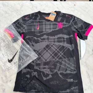  Chelsea fotbollströja Nike - Chelsea fotbollströja från Nike i svart och grått med grafiskt rutigt mönster. Kortärmad modell med rosa och gula detaljer på ärmarna samt broderat lejon på bröstet. Tillverkad i lätt och ventilerande polyester, perfekt för träning eller match.