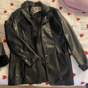 Svart skinnkavaj från Gina Tricot - Snygg svart kavaj i skinnimitation från Gina Tricot med klassisk krage, knappar framtill och tre fickor. Perfekt för dig som vill ha en stilren och trendig look. Fodrad insida och långa ärmar.