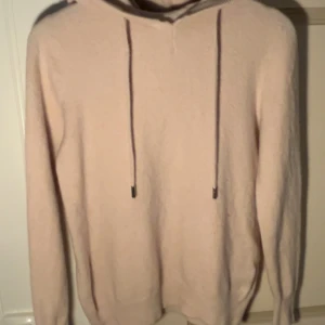 Beige hoodie med huva och snörning - Mysig ljusrosa hoodie med huva och långa ärmar. Tröjan har snörning framtill och är tillverkad i ett mjukt stickat material som känns skönt mot huden. Perfekt för dig som gillar en enkel och chill stil. En liten deffekt vid kragen som smått blivit riven isär men har sytts ihop. Plagget har tagit väl hand om gällande tvätt, bara handtvätt.