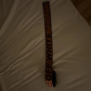 Orange Off-White bälte med svart text - Säljer ett beige bälte från Off-White med tydlig svart text och metallspänne. Bältet har en cool streetwear-vibe och är perfekt för att lyfta din outfit. Texten och detaljerna ger en unik look som sticker ut.