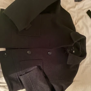 Svart ullrock från 157, XS - Stilren svart rock från 157 i storlek XS. Jackan har klassisk krage, stora framfickor och knäppning med tre knappar framtill. Tillverkad i ullblandning som håller dig varm under kyliga dagar. Perfekt för dig som gillar en clean och tidlös look.