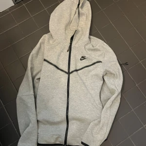 Nike tech - Nike hoodiejacka i ljusgrått med svart dragkedja och svarta detaljer. Jackan har huva, lång ärm och en clean design med Nike-logga på bröstet. Tillverkad i mjuk bomullsmix, perfekt för chill eller träning. Snygg passform och stilren look.