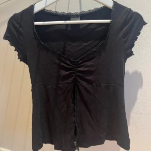 Svart omlottopp med spetsdetaljer - Svart omlottopp med v-ringning och spetskant längs halsringningen. Toppen har korta ärmar med vågig kant, snörning framtill och en öppning i nederdelen. Mjuk och stretchig känsla, perfekt för en trendig look.