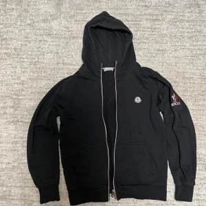 Svart Moncler hoodie med dragkedja - Bra hoodie saken är att den inte går att zippa upp den, men borde vara en enkel fix. Pga så säljer jag den billigare.                                             Svart hoodie från Moncler med broderad logga på bröstet och tryck på ärmen. Tröjan har hel dragkedja med rödvit detalj, två fickor fram och klassisk huva. Tillverkad i mjuk bomull, perfekt för en chill och stilren look.