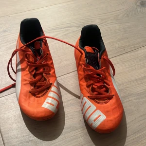 Puma orange fotbollsskor med snörning - Säljer ett par grymt snygga orangea fotbollsskor från Puma med vit logga och detaljer. Skorna har rund tå, platt sula och klassisk snörning. Yttermaterialet är syntet och insidan är svart för extra kontrast. Perfekt för dig som vill sticka ut på planen!