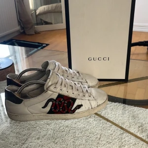  sneakers med orm broderi - Säljer ett par vita sneakers i skinn med broderad röd och svart orm på sidan, gröna och röda ränder samt rund tå och platt sula. Skorna har snörning och klassisk -logga på sulan. Perfekt för dig som vill sticka ut med lyxig streetstyle.