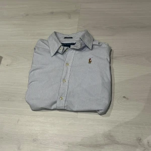 Randig skjorta från Ralph Lauren - Klassisk skjorta från Ralph Lauren i ljusblått och vitt med smala ränder. Skjortan har lång ärm, knappar framtill och ikonisk broderad logga på bröstet. Tillverkad i bomull och har en normal passform. Perfekt för en clean och stilren look.