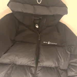 Svart Sail Racing dunjacka. stl M - Svart dunjacka från Sail Racing med huva och dragkedja framtill. Köptes för 4000kr Jackan är fodrad med 500 FP Allied Feather + Down och har en clean, puffad design. Logotypen syns på bröstet och jackan har en modern, oversized look som är perfekt för kalla dagar.